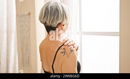 A mature woman with grey hair and a tattoo looks away in a sunlit bedroom, conveying serenity and introspection. Stock Photo