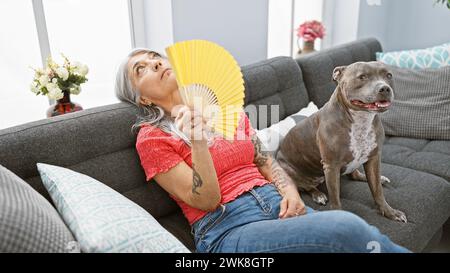 In the heat at home, a mature middle age, grey-haired woman sitting comfortably on the cozy sofa, relaxes fanning air with a hand fan, while her belov Stock Photo