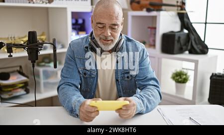 Bald mature man in denim jacket engaging with smartphone in an office setting, showcasing technology use by seniors. Stock Photo