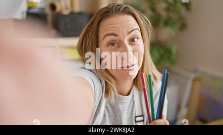 Beautiful blonde young woman artist in apron cheerily showing pencils on video call from her creative art studio Stock Photo