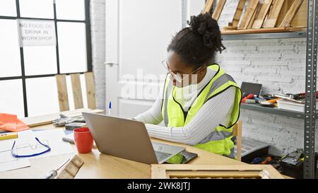 Focused african woman working with laptop in carpentry studio surrounded by tools and wood. Stock Photo