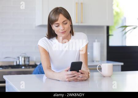 Woman enjoys leisure time with phone and coffee in kitchen. Stock Photo