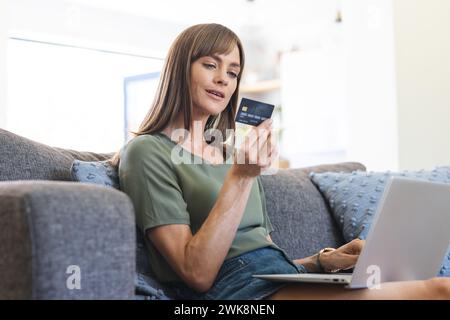 A middle-aged Caucasian woman is focused on her credit card while using a laptop Stock Photo