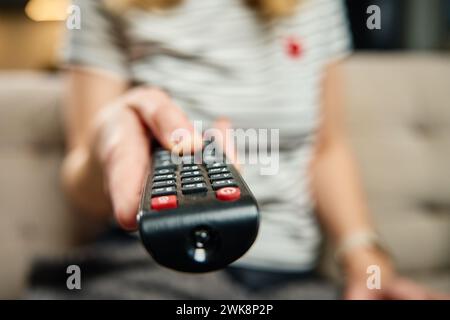 Woman holds TV remote control. Female is sitting on sofa in living room and switching channels on the TV. Concept of entertainment and lazy weekends Stock Photo