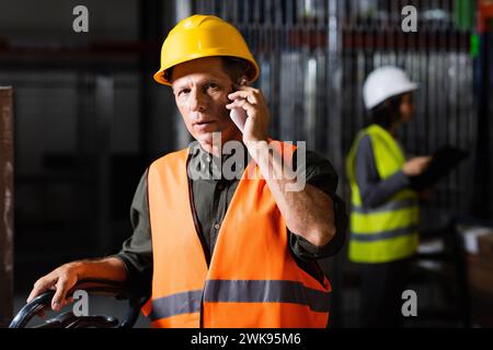 Warehouse supervisor making a phone call on smartphone with a female colleague in the background Stock Photo