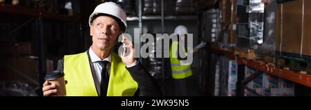 middle aged worker in hard hat and safety vest holding coffee to go while having phone call, banner Stock Photo