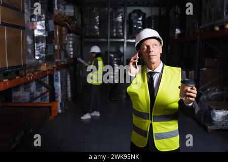 middle aged worker in hard hat and safety vest holding coffee to go while having phone call Stock Photo