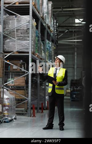 Confident supervisor in safety vest and hard hat inspecting warehouse with clipboard, professional Stock Photo