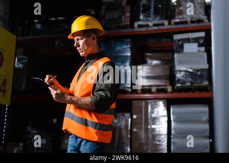 middle aged warehouse supervisor in hard hat taking notes on clipboard while inspecting cargo Stock Photo