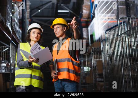 Two warehouse workers discussing logistics, middle aged man pointing near woman with folder Stock Photo