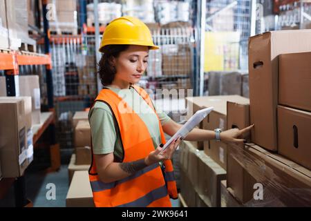 female warehouse worker in safety vest and hard hat holding digital tablet and inspecting cargo Stock Photo
