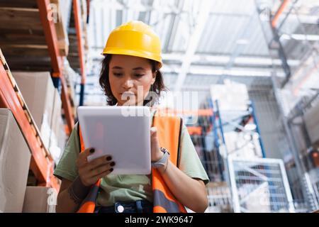 focused female warehouse worker in safety vest and hard hat holding digital tablet near cargo boxes Stock Photo