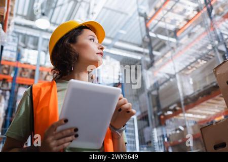 female warehouse worker in safety vest and hard hat holding digital tablet and checking cargo Stock Photo