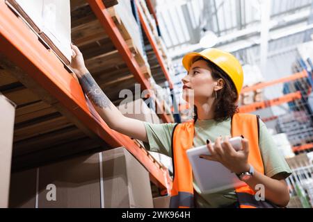 tattooed female warehouse worker in safety vest and hard hat holding digital tablet near cargo boxes Stock Photo