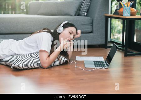 Young asian woman with headphone open mouth yawning while lying on the floor in living room. Studying online at home. Distance learning. Remote work p Stock Photo