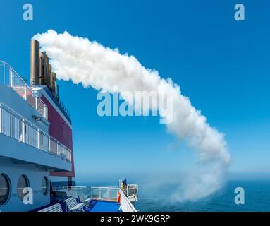 In a composition in blue, white and red, a ferry bound for England trails a dense white cloud of exhaust fumes from its funnels. Stock Photo