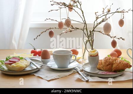 Easter breakfast with egg, bun, coffee, croissant, cheese and sausage, decorated with spring branches and hanging easter eggs in a glass vase on a tab Stock Photo