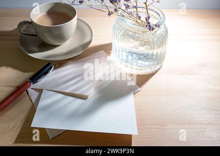Greeting card and envelope from natural recycling paper, pen, coffee cup and a glass vase with sunlight reflections in the morning on a wooden desk, w Stock Photo