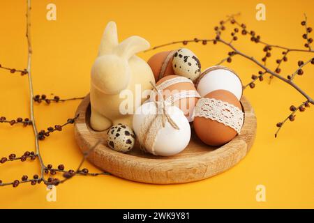 Wooden plate with Easter eggs, bunny figure and branches on yellow background Stock Photo