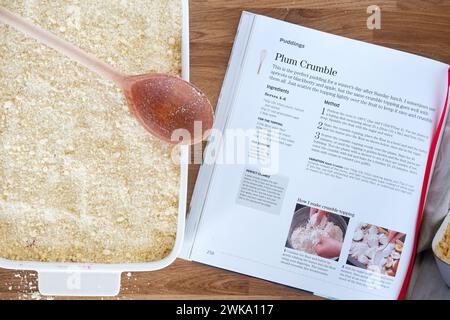 A homemade traditional plum crumble. The desert is made in a large ceramic baking dish and next to the book containing the recipe used to make it. Stock Photo