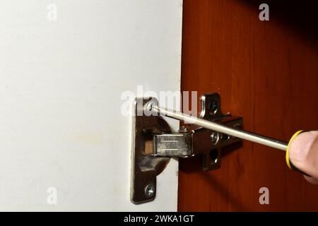 A close-up of a worker's hand, who is busy repairing furniture, unscrews a cabinet door hinge bolt. Stock Photo