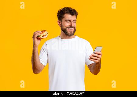 Cheerful young man holding burger using cellphone over yellow background Stock Photo