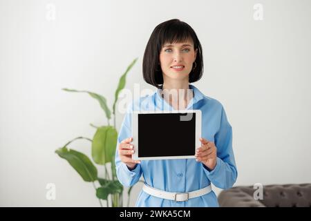 Portrait of young confident modern smiling businesswoman in formal blue shirt shows digital tablet with empty black screen, and looks at camera. Empty Stock Photo