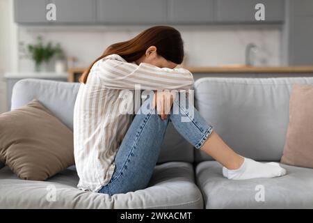 Upset young woman sitting on the sofa with her head down, side view Stock Photo