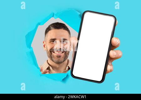 Smiling bearded man looking through a ripped cyan paper, extending a hand to present a smartphone Stock Photo