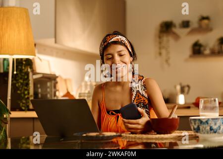 Happy Hispanic businesswoman looking through messages in smartphone while sitting in front of laptop by kitchen table during network Stock Photo