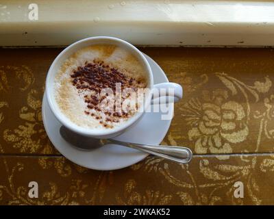 Cappuccino with chocolate sprinkles in a white cup and saucer on an antique table with a decorative pattern. Stock Photo