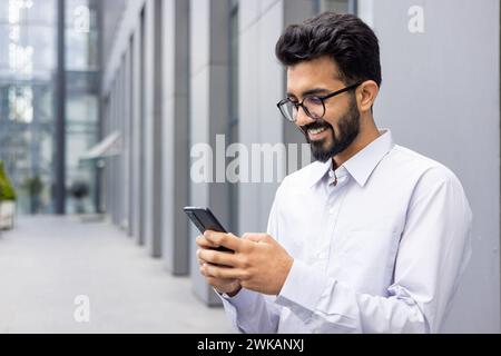 A young businessman from outside an office building uses the phone, a man smiles and browses social networks, a company employee in a shirt with a beard walks around the city. Stock Photo