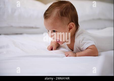 Authentic portrait of a Caucasian cheerful playful little baby boy 4-5 months old in a white bodysuit, lying on his stomach tummy and smiling, trying Stock Photo
