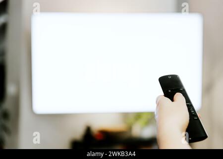 Hand holding remote control pointing to TV. Person watching smart television indoors. White empty mockup screen, copy space. Movie stream service. Video on demand subscription service, platform  Stock Photo
