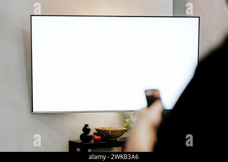 Hand holding remote control pointing to TV. Person watching smart television. Blurred background. White empty mockup screen, copy space. Manage appliances, elements of a smart home control panel Stock Photo