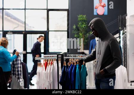 African american robber trying to steal casual wear, looking around to see if someone watching him. Thief robbing clothing store, wearing hood and sunglasses while stealing accessories. Crime concept Stock Photo