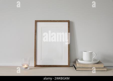 Blank vertical wooden picture frame mock up on wooden table. Modern interior. Cup of coffee on pile of vintage books, burning candle. White wall Stock Photo