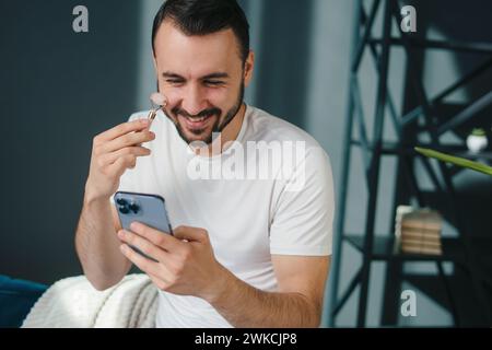 Bearded man using jade face roller for beauty facial massage, holding mobile phone. Concept of natural beauty, skincare, facial expression, anti-aging Stock Photo