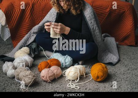 Young woman knitting at her leisure time while sitting on the floor in the evening. Knitting at home. Carefree lady doing knitting work as a hobby Stock Photo