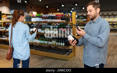 Man and woman customers in liquor store choose wine using mobile phone for reading e-label on wine bottle. Stock Photo