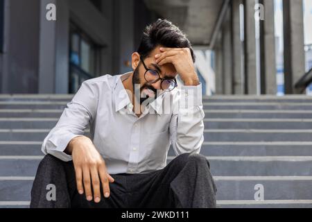 A stressed businessman in despair sits on outdoor steps, expressing worry and frustration, embodying work-related stress. Stock Photo