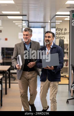 Two mature executives walking together in the office, one with laptop and one with documents.  Stock Photo