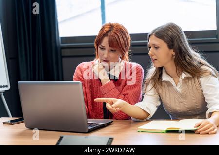 Redhead and brunette working together, discussing a project on a laptop in a bright office.  Stock Photo