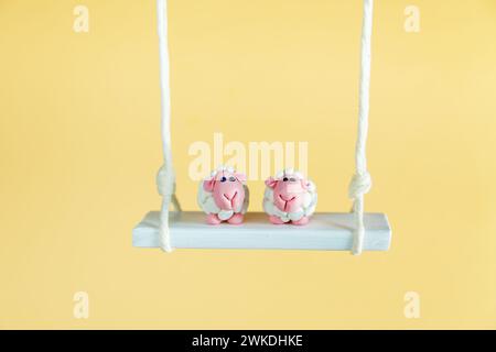 Two cute plasticine sheep are sitting on a swing. Yellow background, copy space. Stock Photo
