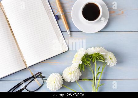 Top view of the desktop. Spiral notebook with an open blank page, glasses, pen, white cup with black coffee, dahlia flowers. Festive office desktop co Stock Photo