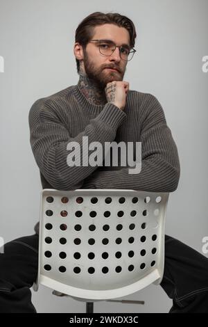 Hipster Handsome Successful Young Guy With A Beard And Hairstyle With A Tattoo In A Fashion Knitted Vintage Sweater With Glasses Eyewear Sits And Pose Stock Photo