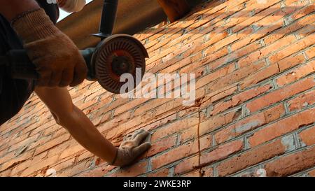 a builder with a grinder and a stone cutting disc stands by a brick wall and applies markings for a future window opening, drawing the outline Stock Photo