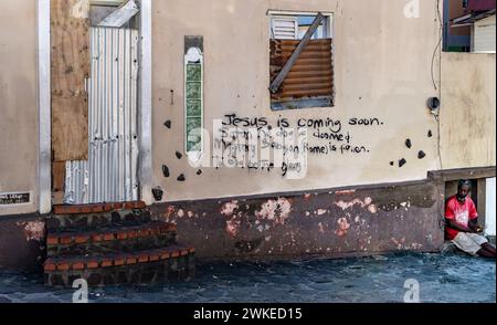 A  beggar sitting in front of a wall with a writing on it about Jesus coming soon and Satan being doomed Stock Photo