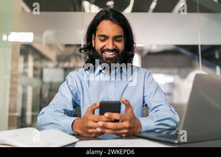 Positive indian guy manager using smartphone, modern office interior Stock Photo