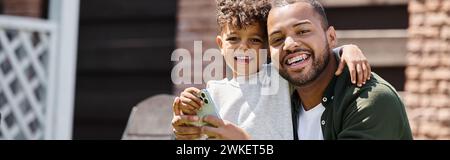 positive african american man in braces and son using smartphone on backyard of house, banner Stock Photo